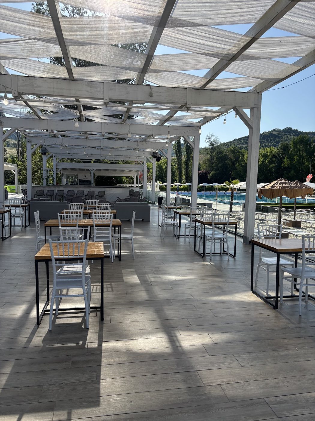 La terrazza di New One: pergolato bianco con teli ombreggianti, tavoli in legno chiaro e sedie bianche, la piscina e le colline sullo sfondo